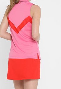 Ärmlös sporttopp i rosa med ett rött chevronmönster och krage; kombinerad med en röd minikjol med sidslitsar. Mjuk tygstruktur.