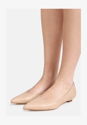 Naakte leren balletplatte schoenen met puntige neus, gedragen op blote voeten, afgebeeld tegen een eenvoudige lichte achtergrond.