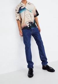 Persona vistiendo jeans azules, zapatos de cuero negros y una camisa de botones de manga corta con un patrón abstracto de nubes en beige, azul y negro.