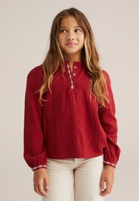 Blusa rossa, con texture, maniche lunghe e scollatura tonda; presenta cuciture decorative bianche lungo il collo e i polsini.