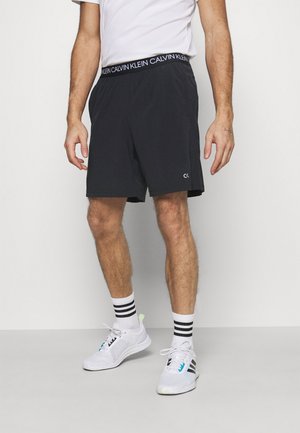 Man draagt zwarte Calvin Klein shorts, wit T-shirt, witte sokken met zwarte strepen en witte sportschoenen, staand tegen een effen achtergrond.