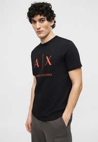 Νεαρός με σγουρά μαλλιά που φοράει μαύρο t-shirt Armani Exchange με κόκκινο λογότυπο και γκρι παντελόνι, στέκεται μπροστά σε απλό φόντο.