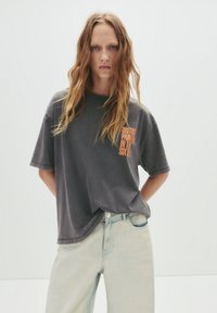 Grå oversized T-shirt med "BRITISH PAINT" grafik i orange på brystet, parret med lyseblå high-waisted denim jeans.