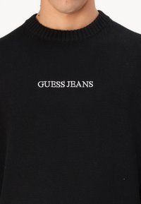 Fekete kötött pulóver bordás nyakkal, a "GUESS JEANS" felirattal hímzéssel fehér színben az elején. Klasszikus környakú szabással rendelkezik.