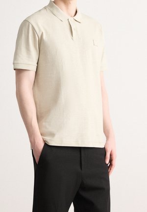 Beige Poloshirt mit kurzen Ärmeln, einem Kragen und einer kleinen Brusttasche. Kombiniert mit schwarzen Hosen, die eine lockere Passform zeigen.