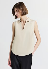 Blusa beige senza maniche con scollo a V e colletto, caratterizzata da una texture liscia e un orlo arrotondato. Indossata con pantaloni neri.