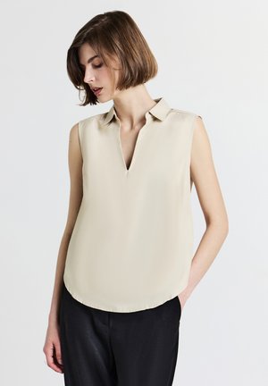 Blusa beige senza maniche con scollo a V e colletto, caratterizzata da una texture liscia e un orlo arrotondato. Indossata con pantaloni neri.