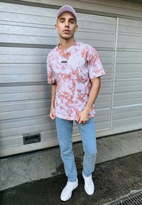 Kurzarm-T-Shirt mit Batikmuster in Pink und Weiß, mit einem rechteckigen Logo, kombiniert mit hellblauen Jeans und weißen Sneakers.