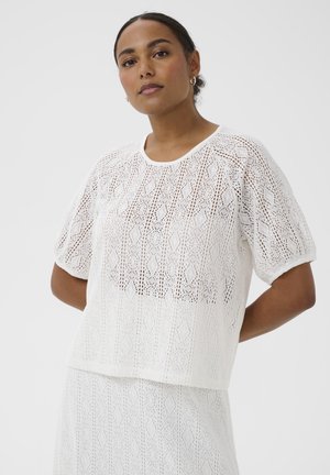 Kobieta o ciemnych włosach związanych w niski kok, ubrana w biały krótki sweter z wzorem i dopasowaną spódnicę na jednolitym tle.