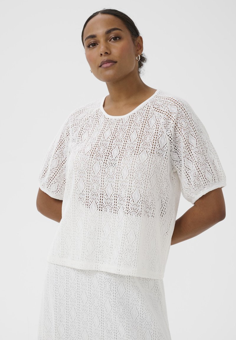 Femme aux cheveux foncés attachés en chignon bas, portant un haut en maille à manches courtes blanc à motifs et une jupe assortie sur un fond uni.