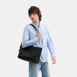 Giovane che indossa una camicia azzurro chiaro e jeans, tiene una borsa a tracolla in pelle nera attraversata sul corpo, guardando di lato.