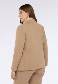 Donna di profilo a sinistra che indossa un blazer e pantaloni in corduroy beige con un dolcevita a coste beige e orecchini a cerchio, su uno sfondo bianco.
