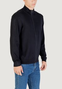 Hombre vestido con un suéter negro de cremallera y cuello alto, junto con jeans azules, frente a un fondo liso.