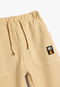 Joggers de tejido de punto waffle en color beige con cinturilla elástica, cordón ajustable y un pequeño parche negro con un logotipo de perro de dibujos animados en el lateral.