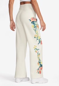 Krämfärgade vida byxor i mjukt material, med ett blommönster i rosa och gult längs vänster sida, med en slät textur.