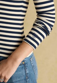 Námornícka a krémová pruhovaná blúzka s dlhým rukávom a ribbovanou textúrou. Má malé logo na manžete. Nosená s modrými džínsami.