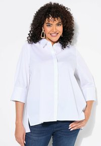 Camicia bianca con bottoni, maniche a tre quarti, spacchi laterali e un fit leggermente loose, realizzata in tessuto morbido. Abbinata a jeans blu scuro.