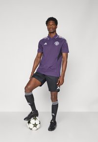 Lilla polo-shirt med sorte accenter, hvide striber og skjold. Kombination med sorte shorts og fodboldstrømper. Sort fodbold ved fødderne.