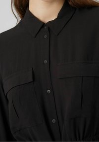 Camicia nera con bottoni, dotata di colletto, due tasche sul petto e vita arricciata. Tessuto liscio, con bottoni neri opachi e cuciture.