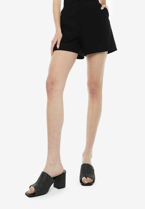 Mule aperti in tessuto nero testurizzato, con punta quadrata e tacco largo, abbinati a pantaloni corti neri. Superficie liscia, design minimal.