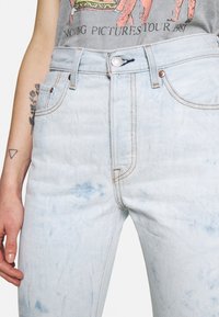 Persona que lleva unos jeans de acid-wash de color azul claro con costuras marrones y una camiseta gráfica gris que muestra un tatuaje parcial en su brazo.