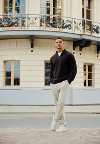 Maglione marrone scuro con zip, pantaloni beige, sneakers bianche. In piedi contro un edificio con un balcone decorativo.