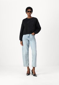 Melnā sudraba džemperis ar teksturētu logotipu, brīvs piegriezums, saskaņots ar gaiši ziliem taisnajiem džinsiem un melnām lakotām augstpapēžu kurpēm. Valda saulesbrilles.