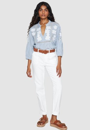 Woman wearing a blue and white striped blouse with white floral embroidery, white pants, brown braided belt, and brown sandals.