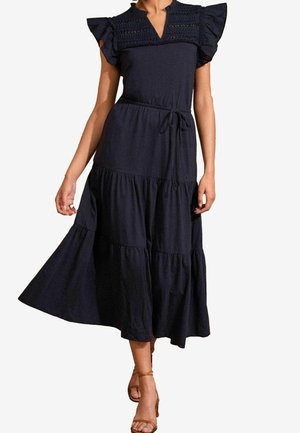 Vrouw draagt een marineblauwe midi-jurk met zwierige mouwen, geplooid lijfje, gelaagde rok en een taillestrik, gecombineerd met bruine sandalen met bandjes.