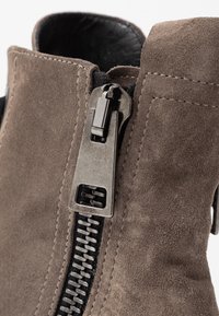Bottines en daim gris avec une fermeture éclair en métal dotée d'une tirette proéminente, d'une doublure noire contrastante et de coutures détaillées le long des coutures.