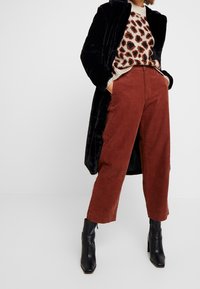 Schwarzer Kunstpelzmantel, beiger Pullover mit braunen Leopardenflecken, rostfarbene Cord-Hose mit weiter Beinform und schwarze Stiefeletten mit quadratischer Spitze.