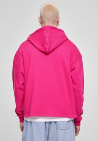Sudadera de forro polar en un vibrante color rosa, con una gran capucha y un corte relajado. Textura suave con puños y dobladillo de canalé, y una pequeña etiqueta en la parte inferior.