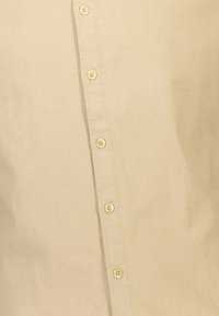 Camicia beige con bottoni, realizzata in un tessuto liscio, dotata di sei bottoni foderati, colletto classico e una finitura del tessuto con un lieve luccichio.