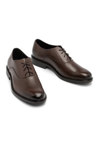 Scarpe in pelle marrone con una texture liscia, punta affusolata e design con lacci. Presentano una suola bassa e robusta nera.