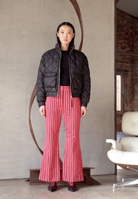 Casaco bomber acolchoado preto, blusa preta ajustada, calças flare listradas em rosa, sapatos pretos com detalhes vermelhos, de pé contra um fundo texturizado.