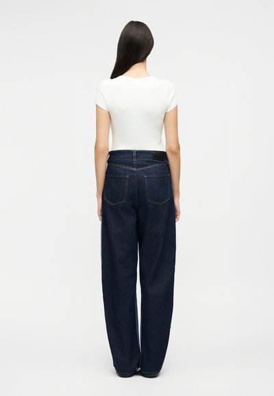 Γυναίκα με μακριά σκούρα μαλλιά που φοράει στενό λευκό κοντομάνικο μπλουζάκι και φαρδιά σκούρα μπλε τζιν, όρθια με την πλάτη στραμμένη προς τον θεατή, σε απλό φόντο.