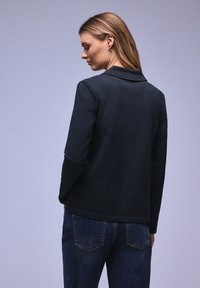 Femme aux cheveux blonds portant un haut à manches longues foncé et un jean bleu, vue de profil sur un fond violet clair uni.