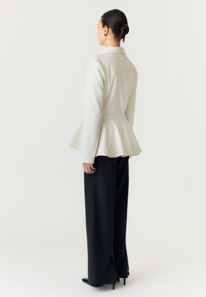 Femme aux cheveux foncés coiffés en chignon, portant une veste blanche à basque et un pantalon noir à jambes larges, debout, dos à un fond uni.
