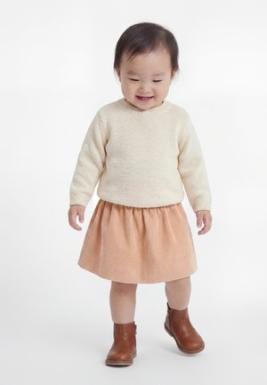 Lillebarn med kort mørkt hår smiler, mens det står iført cream sweater, ferskenfarvet nederdel og brune ankelstøvler mod en hvid baggrund.