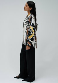 Chemise à manches longues blanche à boutons avec des motifs abstraits noirs et bruns et des accents jaunes, associée à un pantalon noir à jambes larges.