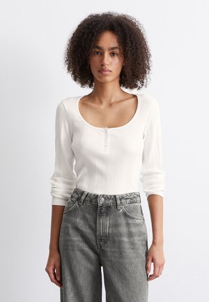 White ribbed long-sleeve top with a scoop neckline, featuring a button placket. Paired with gray high-waisted jeans.