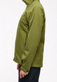 Olivgrön jacka med dragkedja framtill, huva och justerbara ärmslut. Har ett mjukt, vattentåligt tyg och en sidficka.