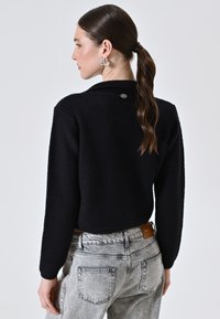 Maglione nero con texture, dal design cropped e maniche lunghe, caratterizzato da un piccolo logo sulla schiena, abbinato a jeans grigi chiari.