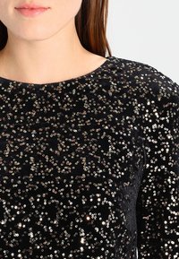 Top en velours noir recouvert de paillettes brillantes dorées et argentées. Doté d'un col rond et d'une surface texturée.