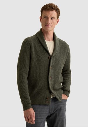 Homme portant un cardigan côtelé vert olive avec col châle et boutons sur une chemise beige, debout avec une main dans la poche sur un fond uni.