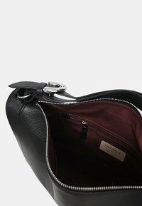 Czarna skórzana torba o teksturowanej powierzchni, z metalowymi elementami w kolorze srebrnym oraz bordowym wnętrzem, wyposażona w zasuwaną kieszeń i detal z etykietą.
