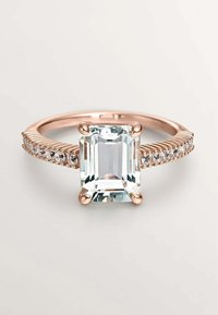 Anillo de gema de corte esmeralda en color transparente, montado en oro rosa. La banda presenta pequeñas piedras de acento a lo largo de un lado con un diseño acanalado.
