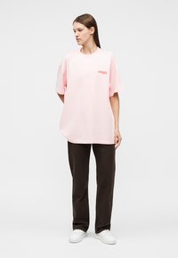 Frau trägt ein lockeres rosa T-Shirt mit kleinem rotem Schriftzug, schwarze Hose und weiße Turnschuhe, steht und blickt nach links vor einem einfarbigen Hintergrund.