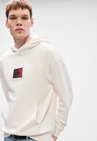 Φούτερ με κουκούλα σε ανοιχτό ροζ ύφασμα, με μαύρη και κόκκινη ορθογώνια ετικέτα λογότυπου που περιέχει το κείμενο "HUGO". Περιλαμβάνει εμπρόσθια τσέπη και ριμπ μανσέτες.