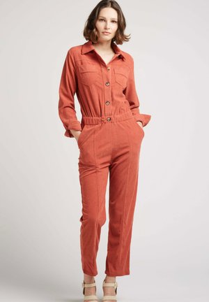 Salopette en velours côtelé couleur rouille avec des manches longues, devant boutonné, deux poches poitrine, taille cintrée et jambes fuselées.